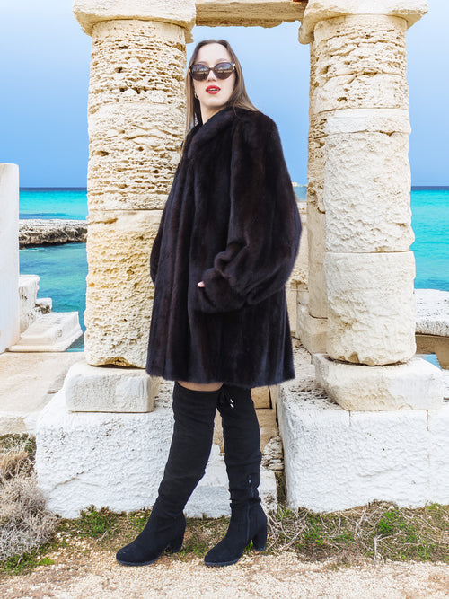 Riviera Noir Black Mink Swing Coat flared open with ocean waves in the background. Statement mink fur coat for luxury winter style.