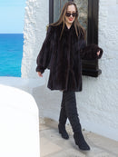 Riviera Noir Black Mink Swing Coat paired with black boots, walking near a white villa. Premium mink outerwear for stylish winter looks.