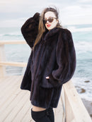 Riviera Noir Black Mink Swing Coat worn on a wooden deck by the ocean. Authentic mink fur coat for upscale cold-weather style.