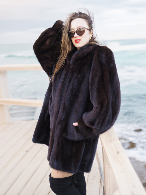 Riviera Noir Black Mink Swing Coat worn on a wooden deck by the ocean. Authentic mink fur coat for upscale cold-weather style.