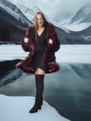 Woman in a fur coat and black dress with knee-high boots standing in front of a snowy mountain landscape.