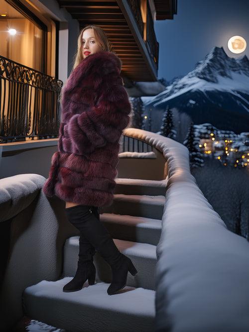 Woman in a fur coat standing on snow-covered steps with a mountainous background at night.