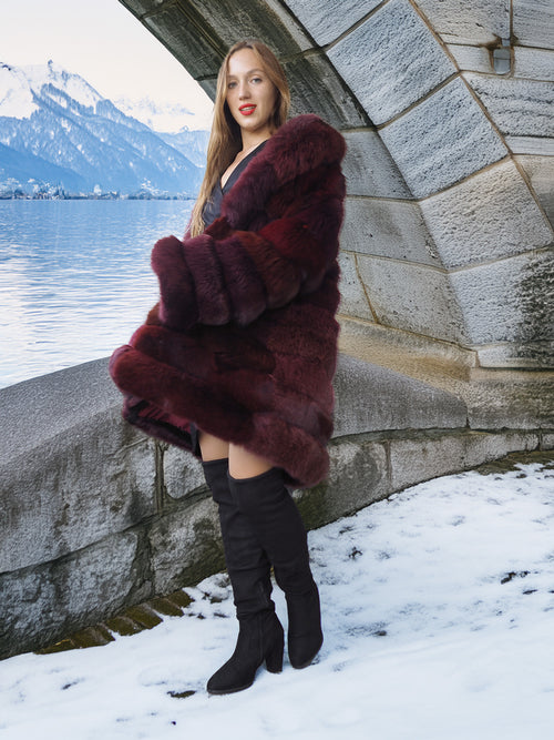 Woman wearing a fur coat standing by a stone wall with a snowy landscape and mountains in the background.