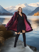 Woman in a black dress and red fur coat standing by a lake with mountains in the background