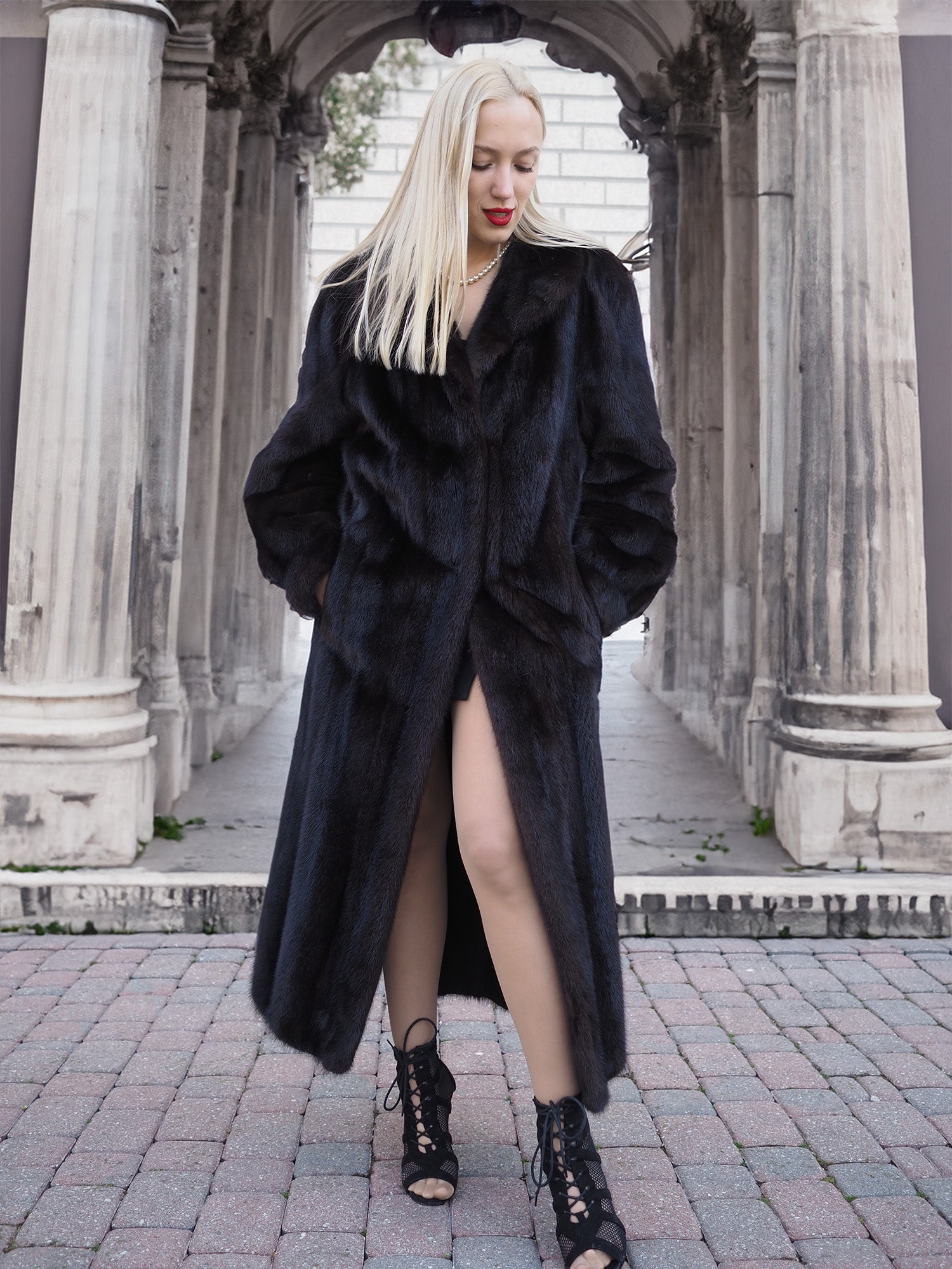 Stylish black fur coat worn by young woman in front of stone columns