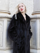 Elegant black mink fur coat with wide lapels displayed on female model against brick wall backdrop.