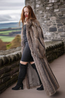 Woman wearing a long fur coat standing in front of a stone wall with a scenic background.