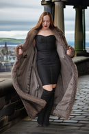 Woman in a long fur coat and black dress standing on a stone pathway with a scenic background.