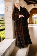 Woman in a long brown fur coat standing against a stone wall with a scenic background.