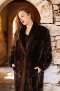 Woman wearing a dark brown fur coat standing against a stone wall.