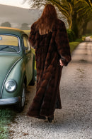 Person in a long brown fur coat walking away from a vintage car on a road.