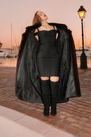 Woman in a black coat and dress standing on a dock at sunset.