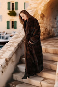 Woman in a long brown fur coat standing on stone steps with a scenic background.