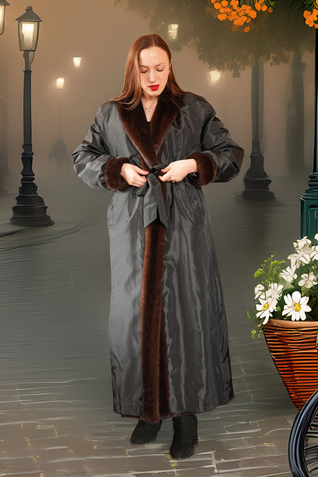 Woman in a long gray coat with brown fur trim standing on a street with lampposts and flowers.