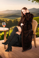 Woman in a fur coat sitting outdoors with a glass of wine, surrounded by scenic mountains.
