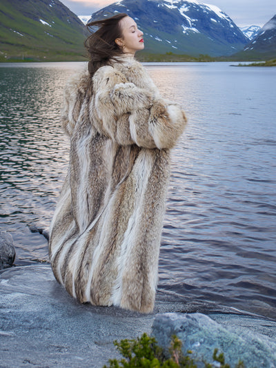 Model in full-length Coyote Fur Coat, enjoying the breeze by a lake with snow-capped mountains in the background.