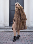 Elegant pastel brown mink fur coat with hooded design, worn by stylish woman against architectural building backdrop.
