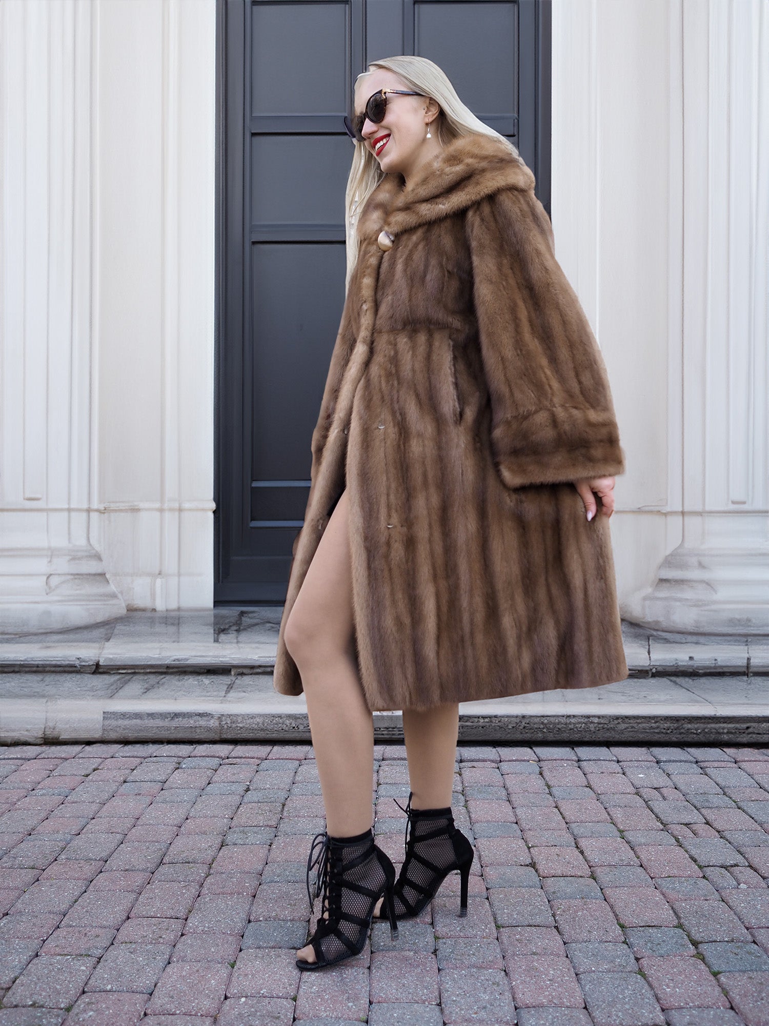 Elegant pastel brown mink fur coat with hooded design, worn by stylish woman against architectural building backdrop.