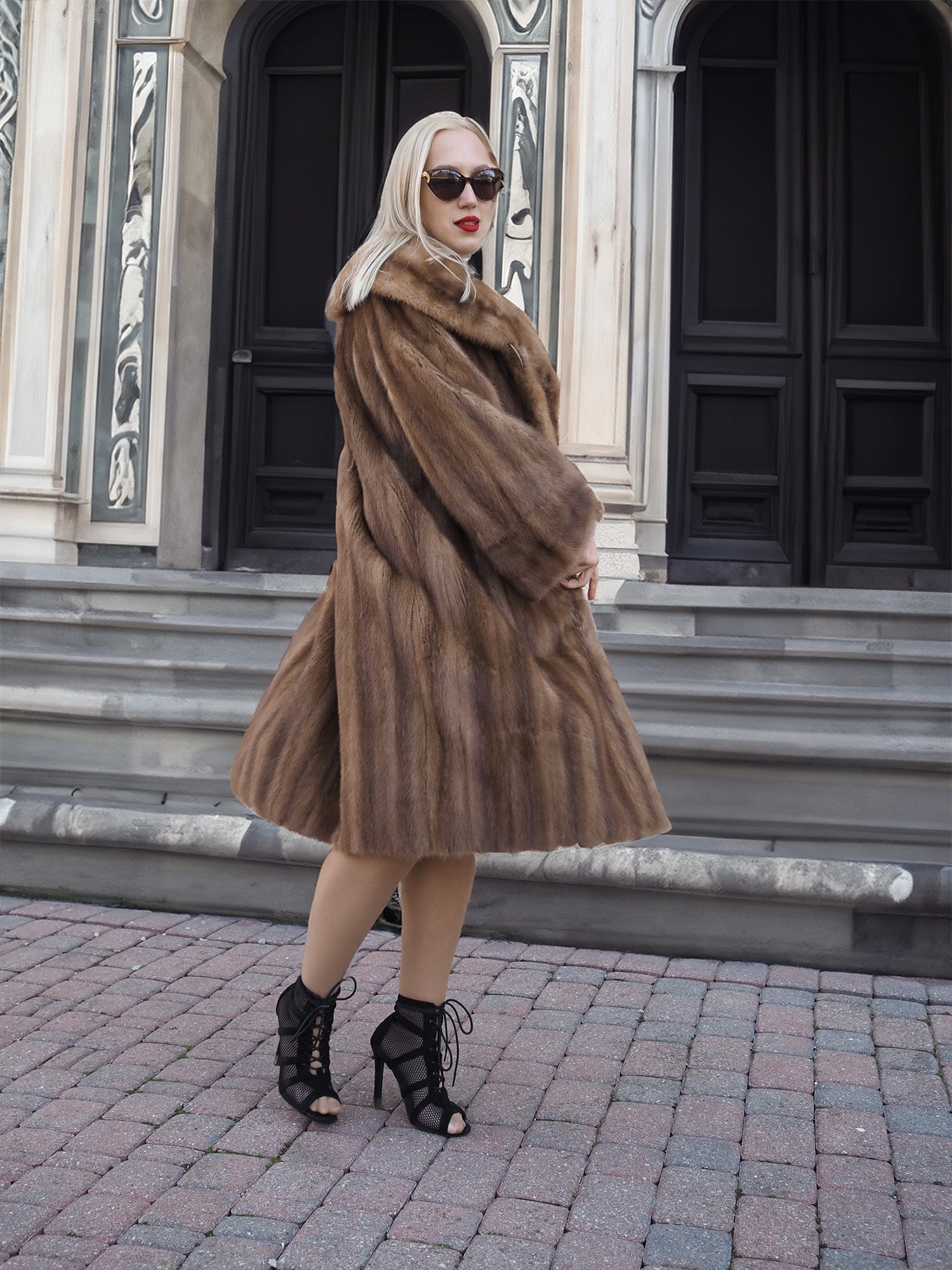Elegant vintage pastel brown mink fur coat, luxurious full-length stroller style, worn by fashionable woman outside architectural building.