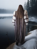 Person wearing a fur coat standing on a snow-covered bank with a lake and trees in the background