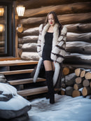 Woman in a fur coat standing in front of a wooden cabin with snow on the ground.
