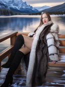 Woman in a fur coat sitting on a wooden bench by a lake with mountains in the background