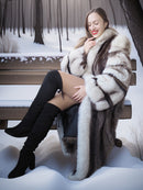 Woman in a fur coat and black boots sitting on a snowy bench.