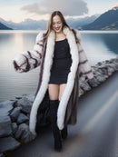 Woman in a fur coat standing by a lake with mountains in the background