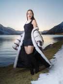 Woman in a long fur coat standing by a lake with mountains in the background