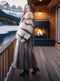 Woman in a fur coat standing on a wooden deck with a mountain and lake view, fireplace in the background.