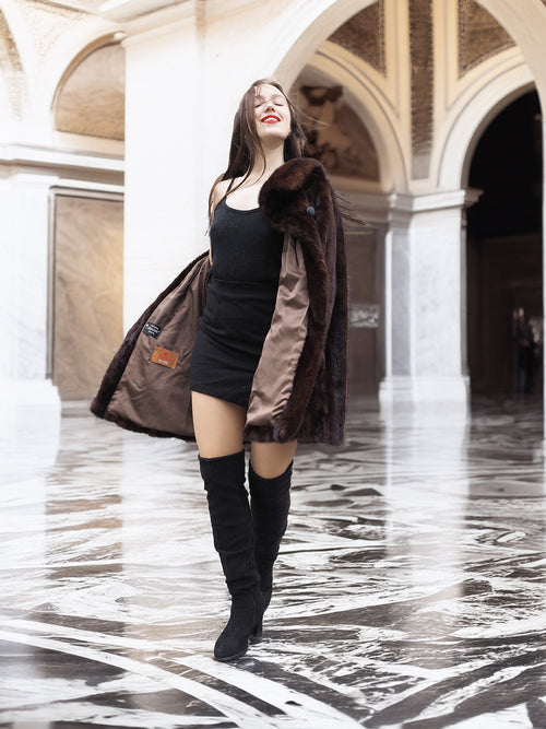 Model twirling in open mink coat, revealing satin lining and elegant black dress in a grand marble interior.