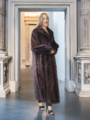 Woman wearing a long brown fur coat standing in an elegant interior setting.