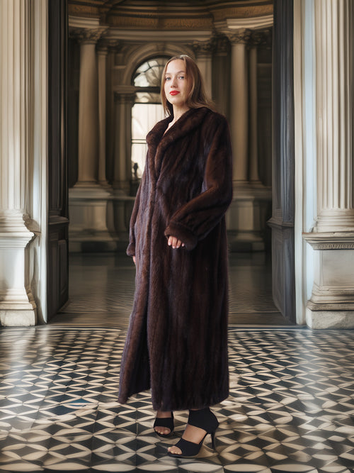 Woman in a long brown fur coat standing in an elegant interior setting with columns and a patterned floor.