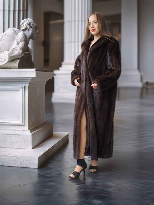 Woman in a long brown fur coat standing in an elegant interior setting with classical architecture.