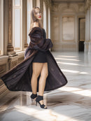 Woman in a long dark coat standing in a sunlit room with classical architecture.