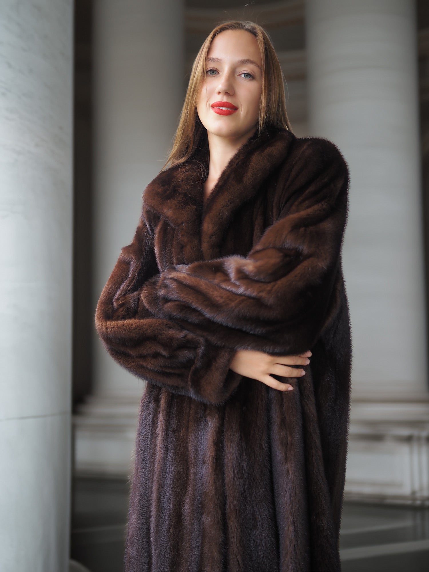 Woman wearing a brown fur coat standing in an indoor setting with columns.