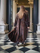 Person wearing a long brown fur coat standing in a grand hallway with columns and a checkered floor.