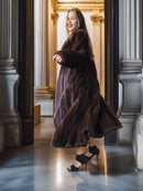 Woman in a long brown fur coat standing in an elegant interior setting.
