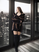 Woman in a fur coat and knee-high boots standing by a large window with a cityscape view.