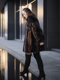 Woman in a dark fur coat and black knee-high boots standing near a reflective surface.