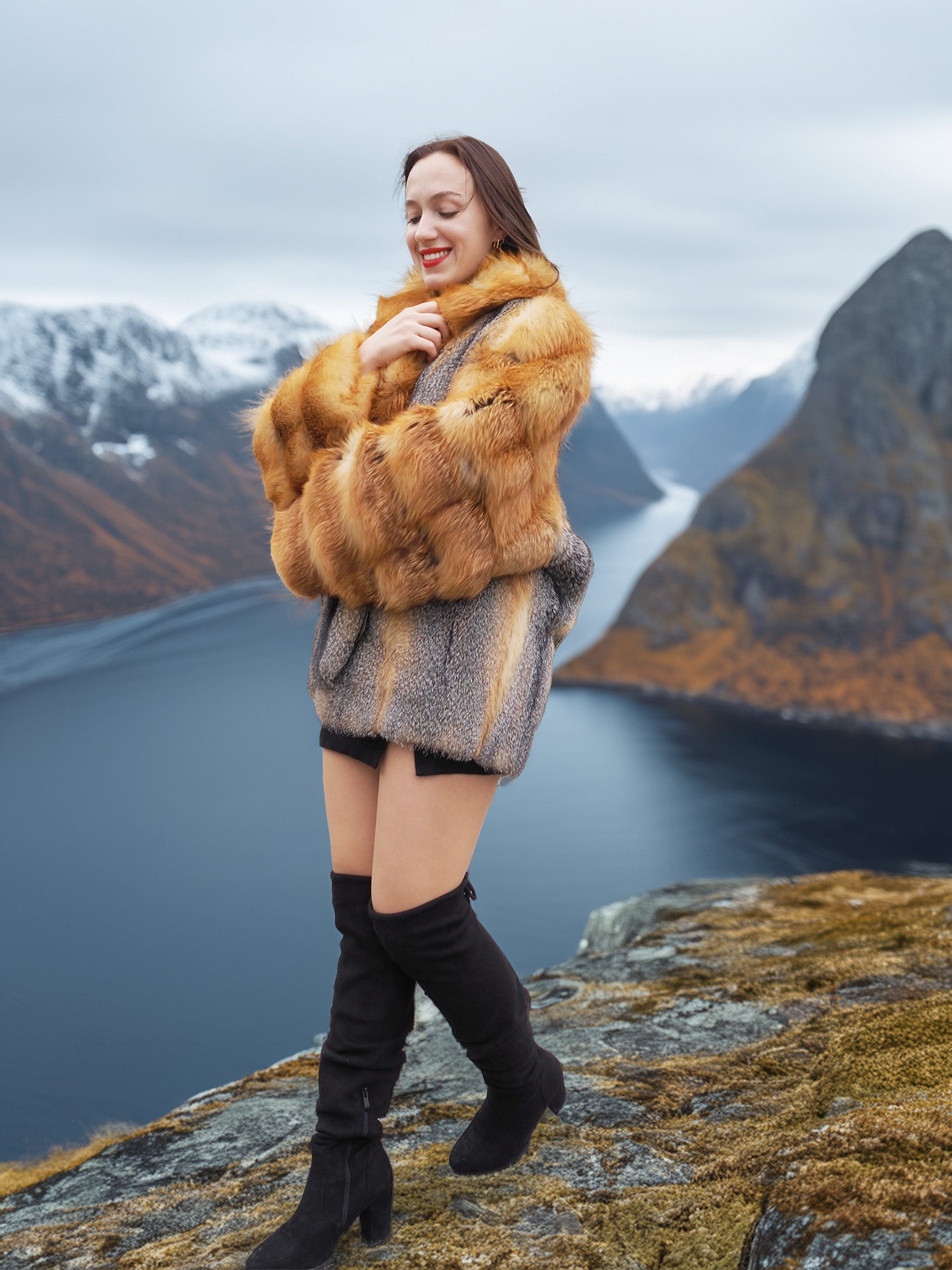 Red and gray fox fur coat by The Wild Muse, modeled with a smile in a dramatic fjord setting, ideal for winter fur fashion lovers.