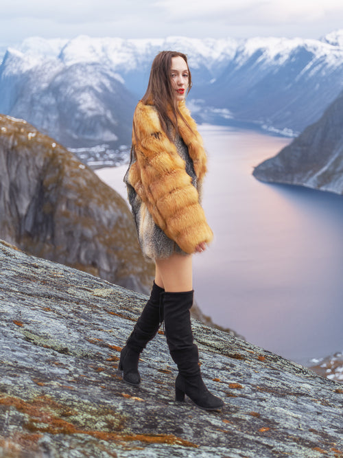 The Wild Muse fox fur coat in red and gray hues, worn while walking along a scenic mountain ridge, ideal for winter outerwear.