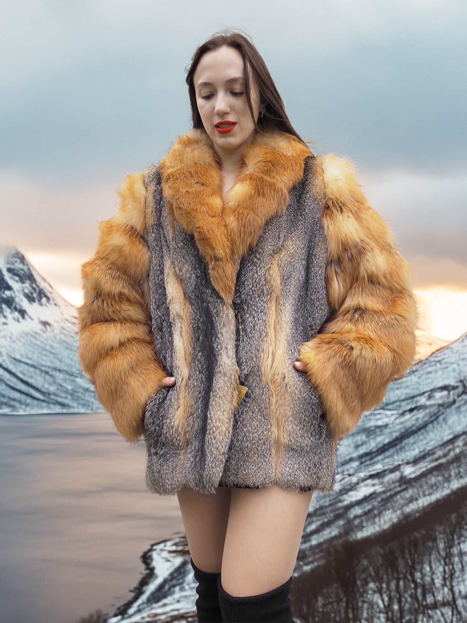 Red gray fox fur coat by The Wild Muse, styled with hands in pockets against a snowy mountain backdrop for a luxury winter look.