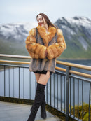 Luxury fox fur coat in red and gray tones by The Wild Muse, modeled on a scenic balcony overlooking snowy mountains and water.