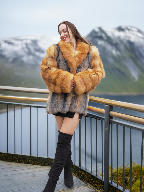 Luxury fox fur coat in red and gray tones by The Wild Muse, modeled on a scenic balcony overlooking snowy mountains and water.