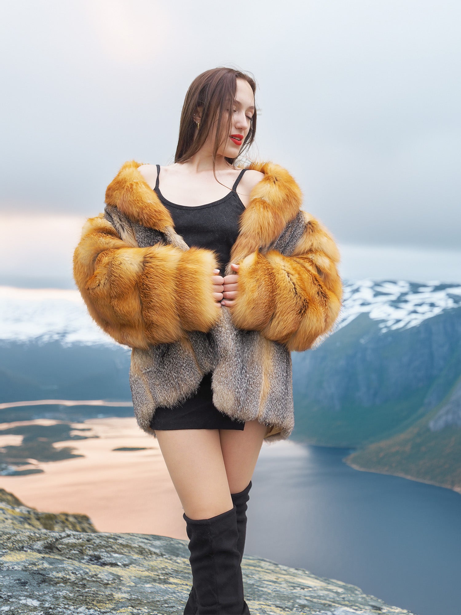 The Wild Muse red gray fox fur coat, styled with a black dress in a snowy fjord setting, showcasing its plush texture and elegance.