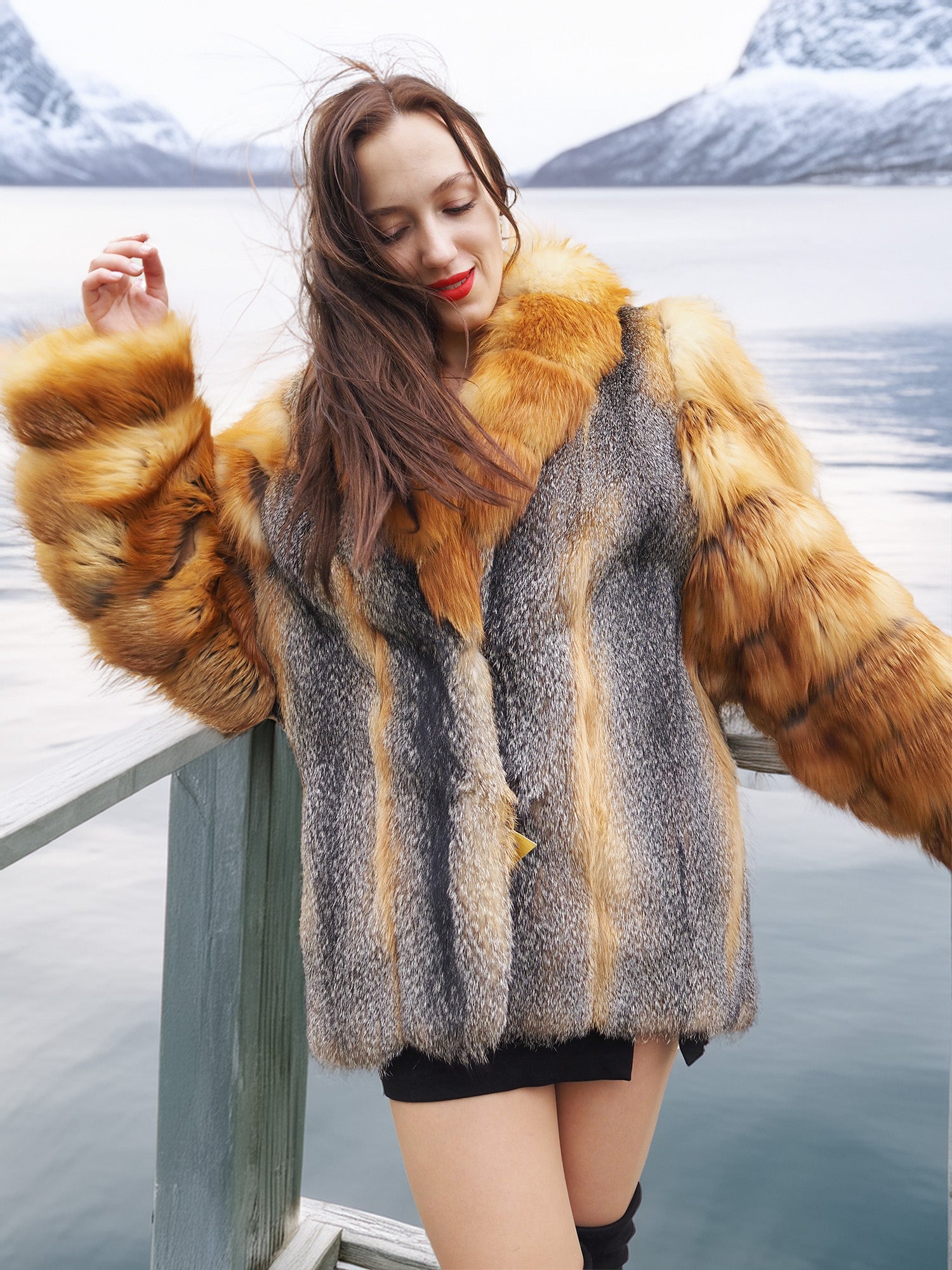 The Wild Muse luxury fur coat in natural red and gray fox, worn on a lakeside deck, showcasing its plush texture and elegant silhouette.