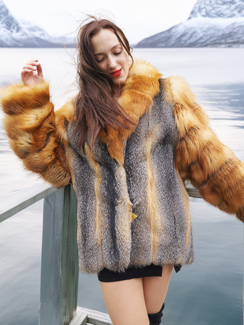 The Wild Muse luxury fur coat in natural red and gray fox, worn on a lakeside deck, showcasing its plush texture and elegant silhouette.