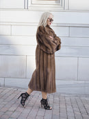 Elegant pastel brown mink fur coat with silk lining, showcased on a blonde-haired woman in high heels against a white brick wall.