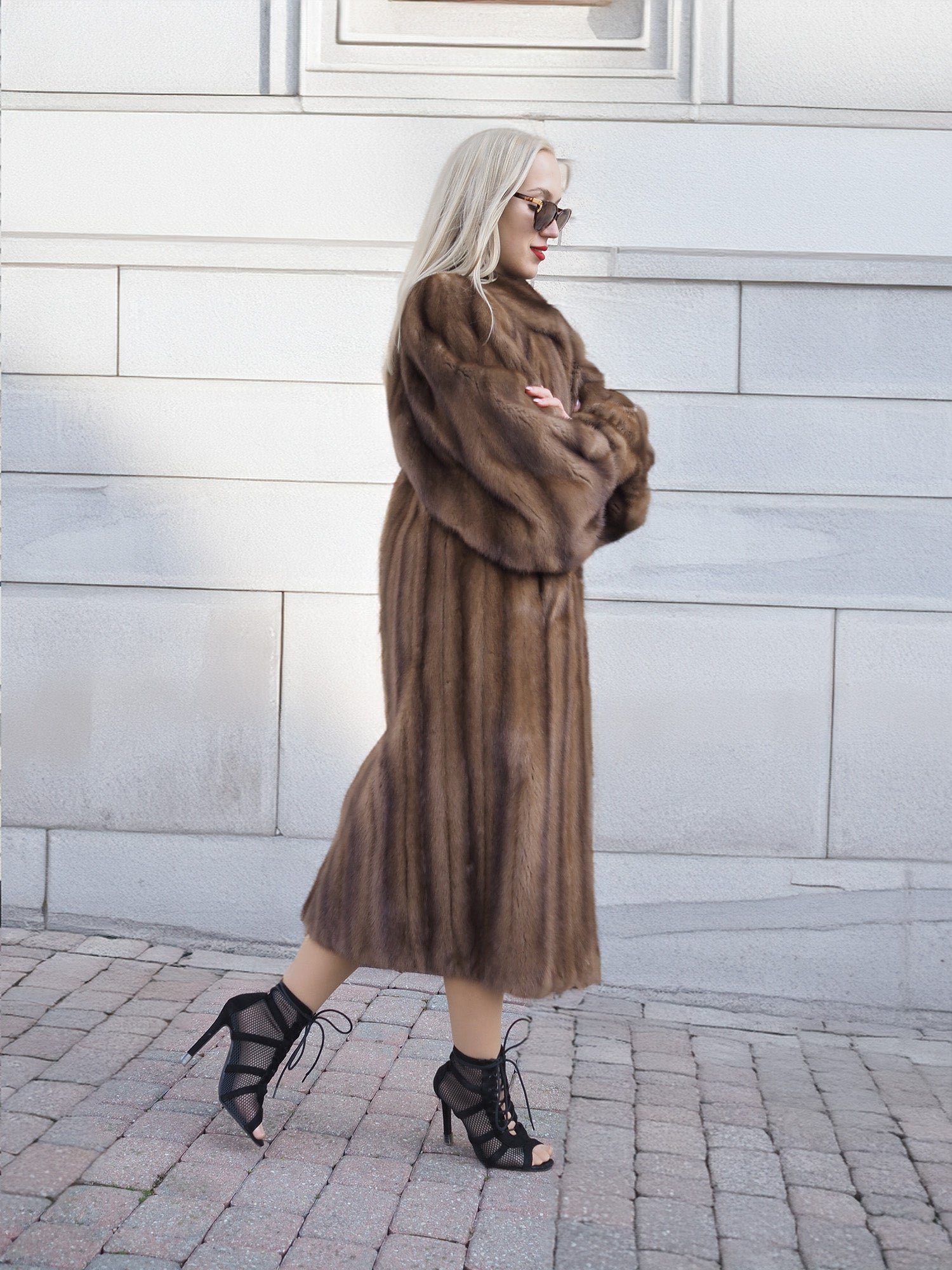 Elegant pastel brown mink fur coat with silk lining, showcased on a blonde-haired woman in high heels against a white brick wall.